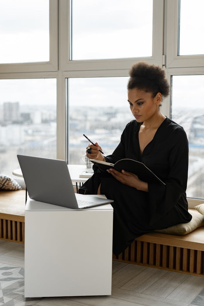 About African American woman studying online on a laptop by a large window in a modern setting.