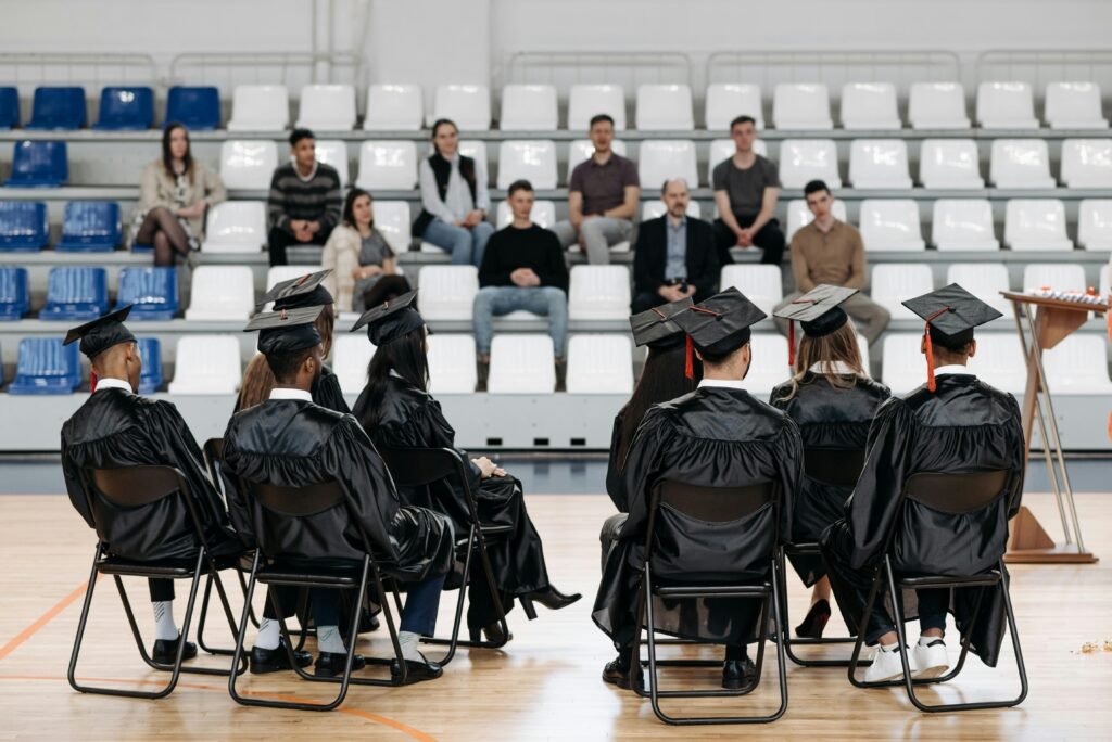 Graduates in gowns sit in a gymnasium with an audience in the bleachers, capturing a special moment.